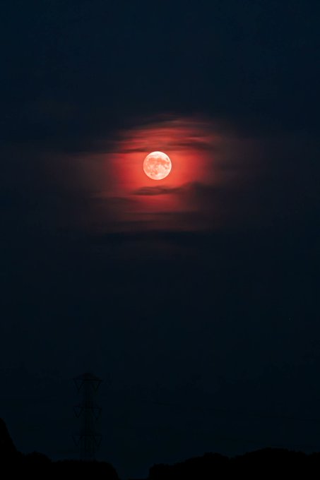 A stunning photograph of a red full moon glowing through dark clouds above a silhouetted landscape with a power line tower.