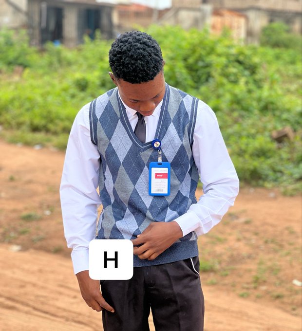 A young man wearing a school uniform with a blue argyle sweater vest, white shirt, and dark tie stands on a dirt path looking down at his ID badge, with a white 'H' icon overlaid on the image.