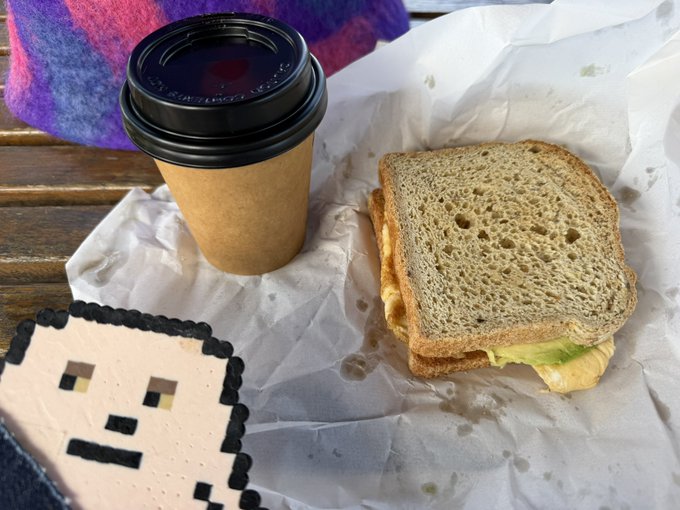 A takeaway meal consisting of a sandwich with lettuce on wheat bread and a coffee cup in a brown sleeve, served on white paper on a wooden surface, next to a pixel-art character sticker.