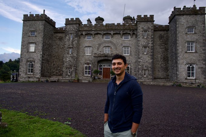A smiling young man in a blue hoodie stands in the gravel courtyard of a large, grey stone castle.