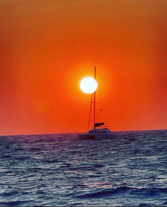 A lone sailboat floats on rippling ocean waters, silhouetted against a fiery orange sunset with the sun glowing brilliantly near the horizon. The serene, dreamy scene captures the essence of the post's reference to "the mulla," evoking a peaceful, aspirational moment on the open sea.