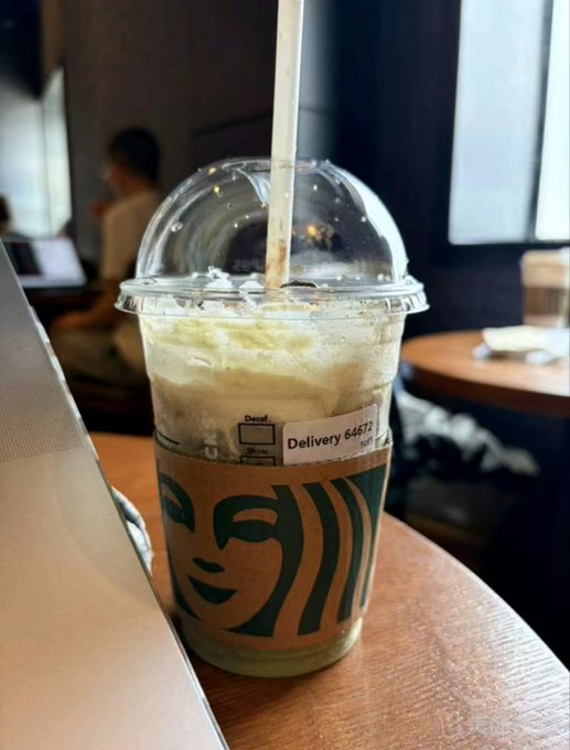 A Starbucks iced drink in a clear plastic cup with a domed lid and straw sits on a wooden table in a cafe setting.