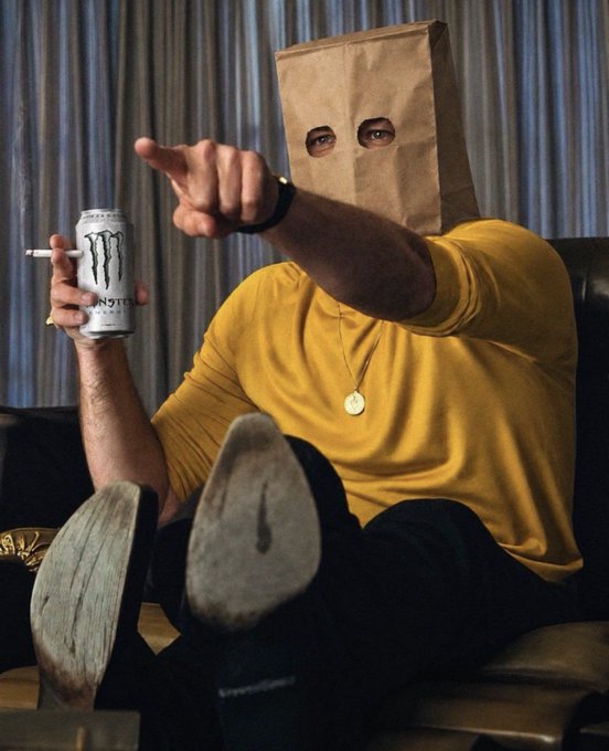 A person wearing a brown paper bag over their head with eye holes cut out, dressed in a yellow turtleneck, pointing at the camera while holding a Monster Energy can and a cigarette.