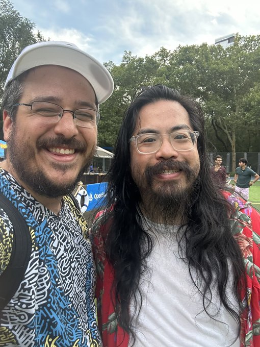 A selfie of two smiling men outdoors; one on the left wears a white cap and glasses, while the other on the right has long dark hair and clear glasses.