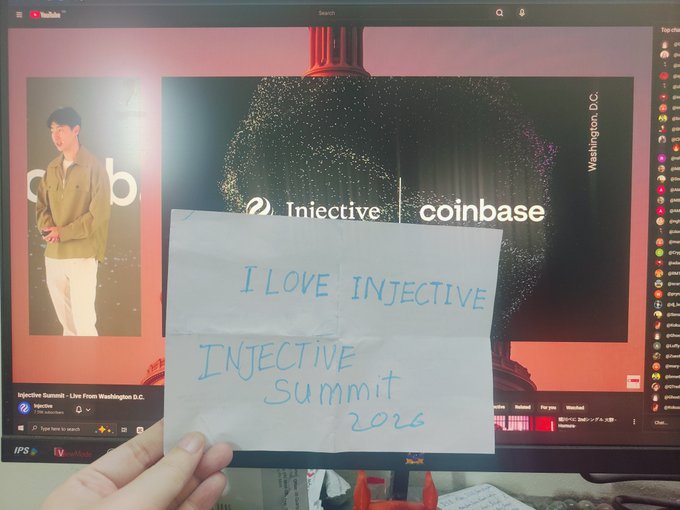 Photo of a hand holding a handwritten note 'I LOVE INJECTIVE / INJECTIVE SUMMIT 2026' in front of a computer monitor displaying an Injective and Coinbase live stream from Washington D.C.