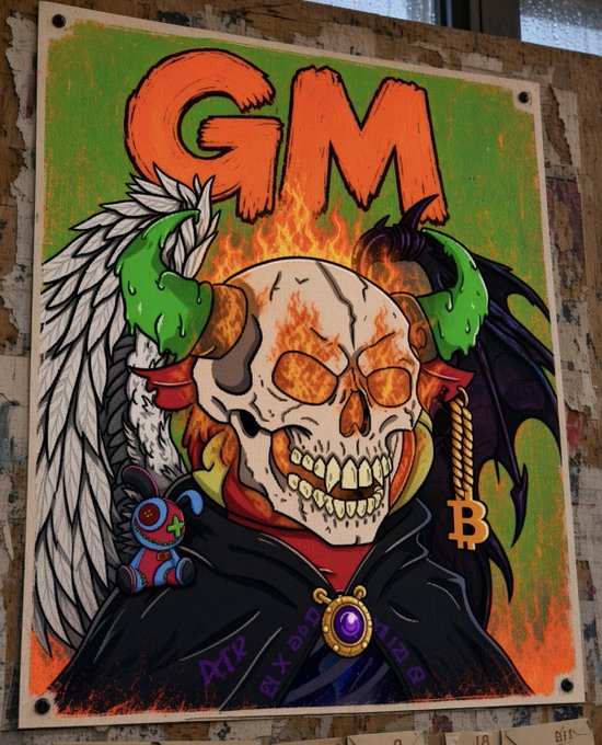 A printed poster featuring a flaming skull in a hooded cloak between white angel and purple demon wings, with the text 'GM' at the top, a Bitcoin symbol, a small rabbit plushie, and 'APE' written at the bottom.