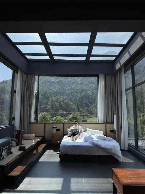 A person sits on a bed reading inside a modern bedroom with large glass windows and a glass ceiling, overlooking a lush, forested mountain valley.