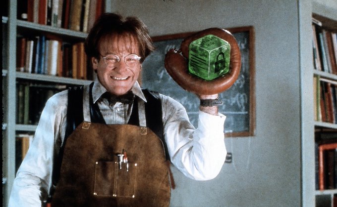 A smiling man wearing glasses and a leather apron holding up a baseball glove with a green velvet box, standing in a library with bookshelves.