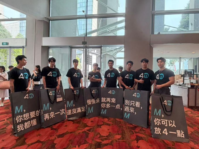 A group of eight young male models wearing matching black "4M" t-shirts stand in a line at what appears to be a hotel or convention center, each holding large gift bags featuring the 4M exchange logo and Chinese promotional slogans like "慢慢来" (take it slow), "别只看" (don't just watch), and "你可以放4一點" (you can relax a bit). The promotional display aligns with the post mocking 4M exchange for their marketing stunt, which has drawn more attention to the handsome models than to the cryptocurrency exchange itself.