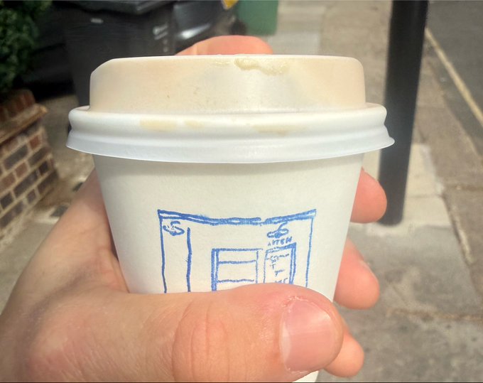 A close-up of a hand holding a white paper coffee cup with a blue line drawing of a storefront on it, taken on a sidewalk.