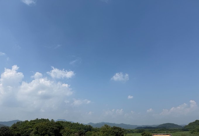 A wide landscape view of a bright blue sky with scattered white clouds above a tree line and distant hills on a sunny day.