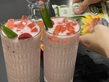 Two tall glasses of pink strawberry milkshakes or smoothies garnished with fresh strawberry slices and green leaves, with someone holding a bottle in the background.