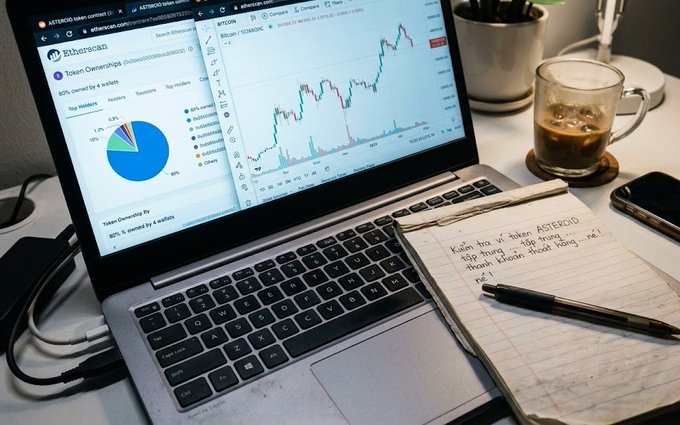 A laptop displaying Etherscan token ownership statistics and a Bitcoin price chart, next to a notebook with handwritten notes and a coffee cup.