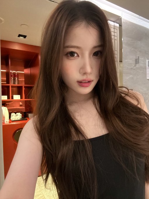 A young woman with long, wavy brown hair poses for a selfie in what appears to be a café or restaurant setting, wearing a black sleeveless top. The image showcases a polished, glamorous look fitting for a brand promotional post tagged with hashtags related to USPEER and SEOYU.