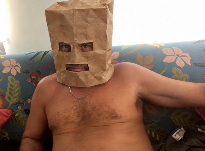 A shirtless man wearing a brown paper bag over his head sits on a floral-patterned blue couch, illustrating a joke about Hunter Biden reportedly canceling his "$LAPTOP" token launch in favor of backing "$ZCAT" due to newfound concerns about privacy—humorously depicted by him covering his face to hide his identity.