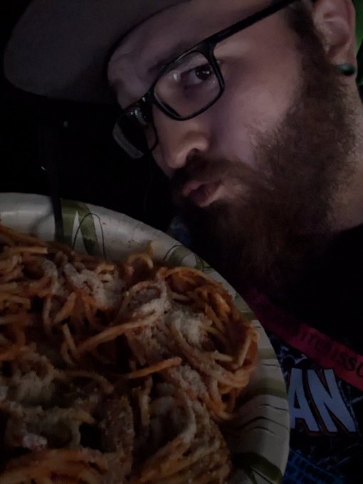 A close-up selfie of a bearded man with glasses and a cap staring intensely at a paper plate piled high with fried food in a dark setting.