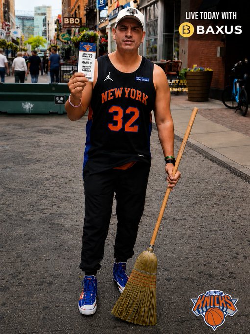The image features a man standing on a street holding a broom, wearing a black jersey with the number 32. The background shows a city street scene with people walking by. There are also two bicycles visible in the picture. The main focus of the image is the man and his broom, which seems to be related to an event or promotion involving basketball fans.