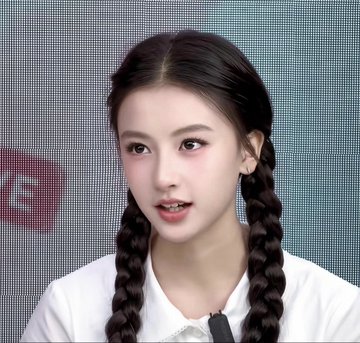 A young woman with dark hair styled in two braids, wearing a white top, speaking with a microphone visible at the bottom of the frame.