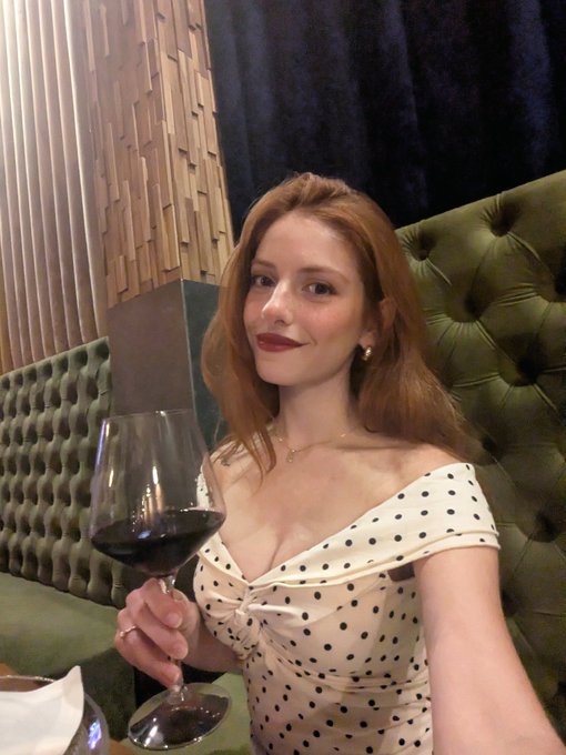 A woman with long red hair wearing a white off-shoulder polka dot dress, holding a glass of red wine while seated on a green tufted leather banquette in a restaurant setting.