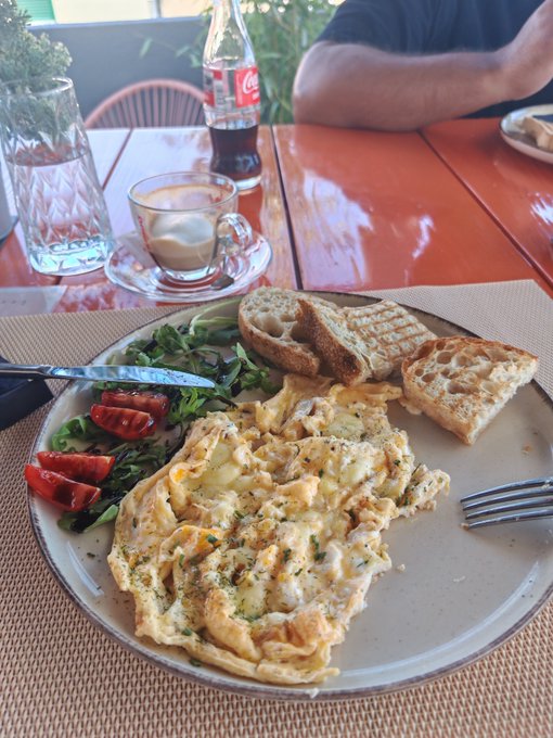 A breakfast plate with scrambled eggs, toast, and a side salad sits on a table next to a Coca-Cola bottle and coffee, with a person seated across the table.