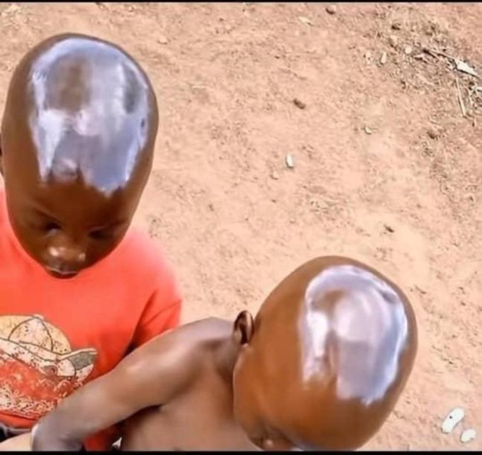 Photo of two young children with shiny heads of hair, one wearing an orange shirt, standing on dry dirt ground.