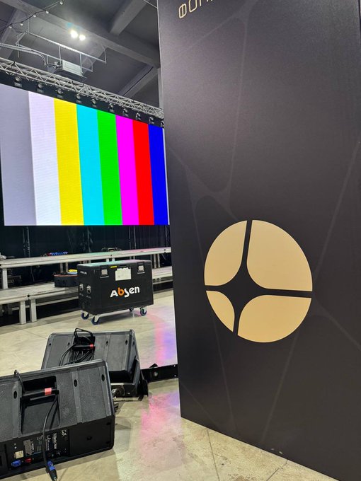 Behind-the-scenes view of a production setup with a large LED screen displaying color bars and a banner featuring a gold circular logo.