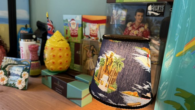A shelf filled with Hawaiian-themed souvenirs, including a yellow ceramic pineapple, a lampshade printed with a Hawaiian scene, Elvis memorabilia, and boxes of local products.