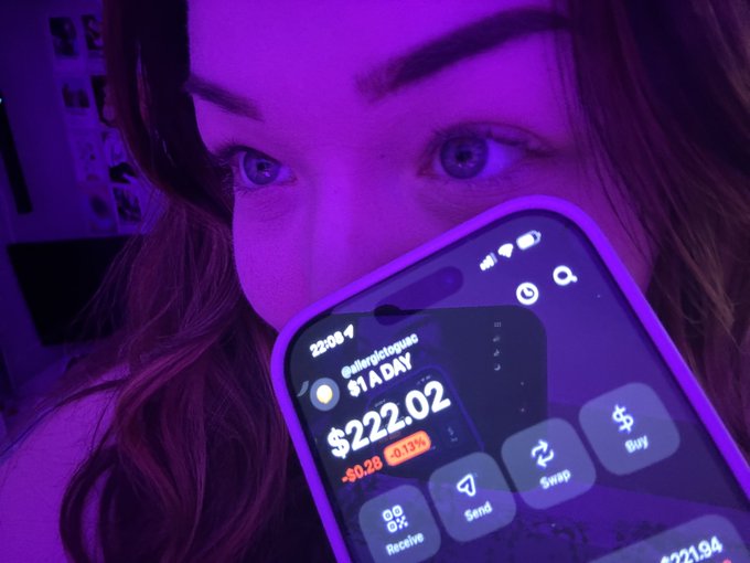 A close-up photo bathed in purple light of a woman holding a smartphone displaying the 'Abstractedgus' cryptocurrency widget, showing a $1 A DAY reward, a $222.02 balance, and a -$0.28 change, with Swap, Send, and Buy buttons visible.