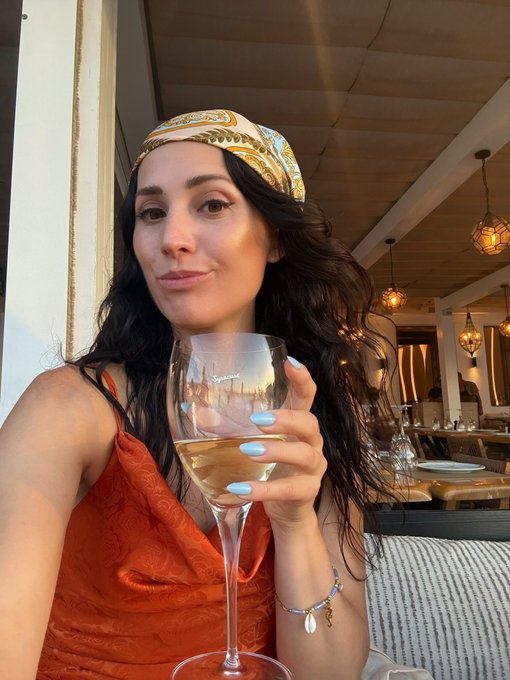 A woman with long dark wavy hair, wearing a patterned silk headband and an orange slip dress, smiles softly while holding a glass of white wine at an upscale restaurant with pendant lighting. Her blue manicure and shell charm bracelet complement the relaxed, golden-hour atmosphere that reflects the post's sentiment of enjoying a well-deserved break from work at Zero Hash.