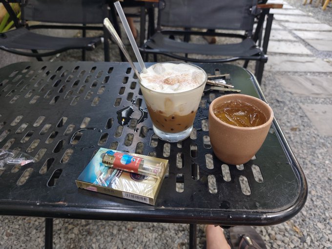 A glass of iced coffee with whipped cream and cinnamon, a brown ceramic cup of coffee, and a pack of Jet cigarettes rest on a black metal outdoor table, suggesting a casual afternoon café break before heading out for drinks.