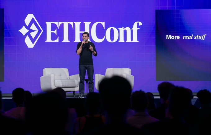 A man speaks on stage at the ETHConf conference, standing between two white armchairs in front of a large purple backdrop displaying 'ETHConf' and 'More real stuff'.