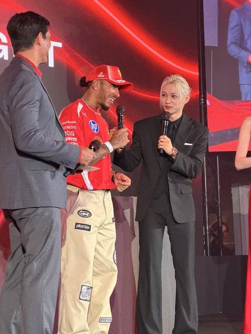 Lewis Hamilton, wearing a red Ferrari racing suit with sponsor logos like HP and Puma, shares a smile on stage alongside a blond-haired man in a black suit, both holding microphones during what appears to be a promotional event.