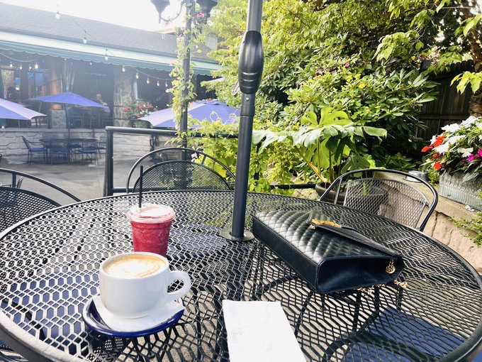 An outdoor cafe table holds a latte, a berry smoothie, a black wallet, and a receipt, with lush greenery and string lights in the background.
