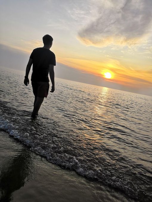 A person in dark clothing wades through shallow ocean water at sunset, with the golden sun reflecting off the calm waves.