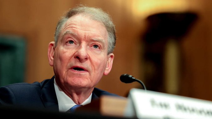 An older man in a dark suit and blue tie speaks into a microphone during what appears to be a formal hearing or press conference.
