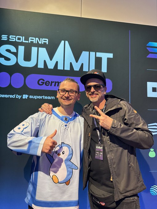 Two men posing at the Solana Summit Germany event — one in a light blue hockey jersey giving a thumbs up, the other in a black leather jacket and sunglasses making a peace sign — in front of a 'SUMMIT' banner powered by superteam.