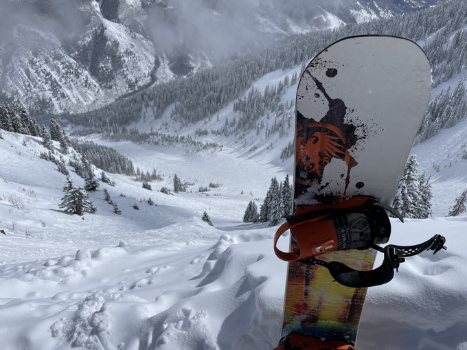 A snowboard with orange and black bindings is stuck vertically in fresh powder snow, overlooking a vast snowy mountain valley dotted with evergreen trees.