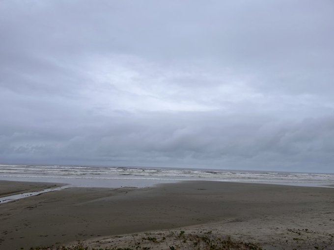 A wide view of a sandy beach under an overcast gray sky, with waves crashing in the distance and small pools of water on the wet sand in the foreground.