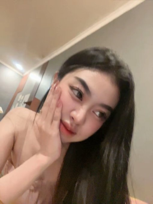 A close-up selfie of a young woman with long dark hair, wearing a pink top, with her hand raised near her face in an indoor setting.
