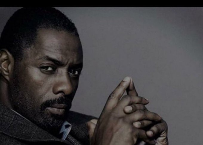 A close-up portrait of Idris Elba, appearing to be in character, with his hands clasped together and an intense expression against a gray background.