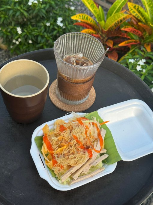 An overhead view of a meal on a black tray outdoors, featuring a styrofoam container with vermicelli noodles, vegetables, and sliced meat on a banana leaf, alongside a cup of tea and a glass of iced coffee.