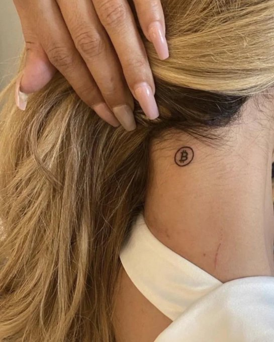 A close-up photo showing a woman's neck behind her ear with a small Bitcoin (₿) logo tattoo, with her hand holding back her blonde hair.