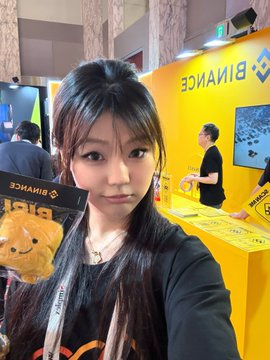 A young woman with long dark hair posing for a selfie at what appears to be a Binance event, holding a small teddy bear plush.