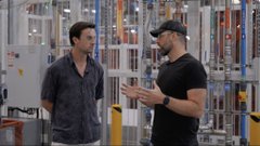 Two men standing in what appears to be a warehouse or store with tall shelving units in the background, one wearing a dark shirt and the other in a black cap and shirt.