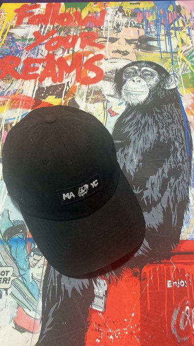 A black baseball cap with a 'MA YC' patch rests on a colorful poster featuring a chimpanzee illustration and the text 'FOLLOW YOUR DREAMS'.