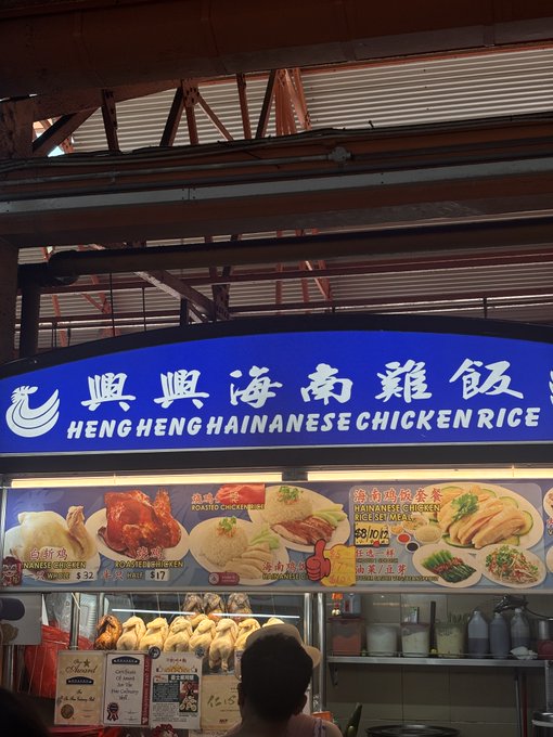 A hawker stall sign reading 'Heng Heng Hainanese Chicken Rice' (興興海南雞飯) displaying various menu items including white chicken, roasted chicken, and rice sets with prices.