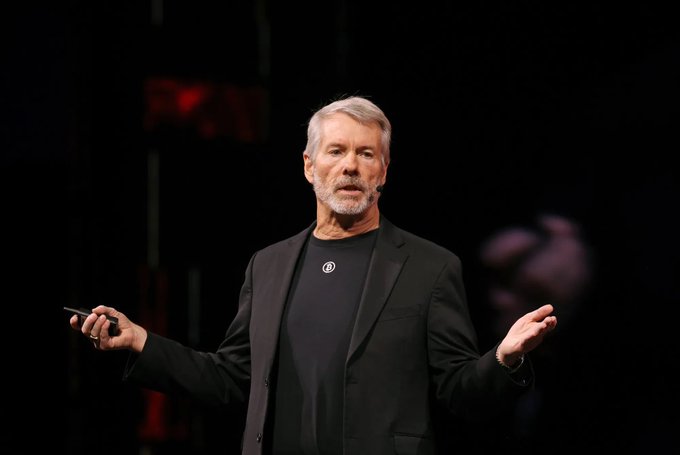 Michael Saylor, executive chairman of Strategy (formerly MicroStrategy), speaks at a conference, gesturing with a clicker, as the post highlights his company's recent Bitcoin sell-offs at a loss after years of promising to never sell.