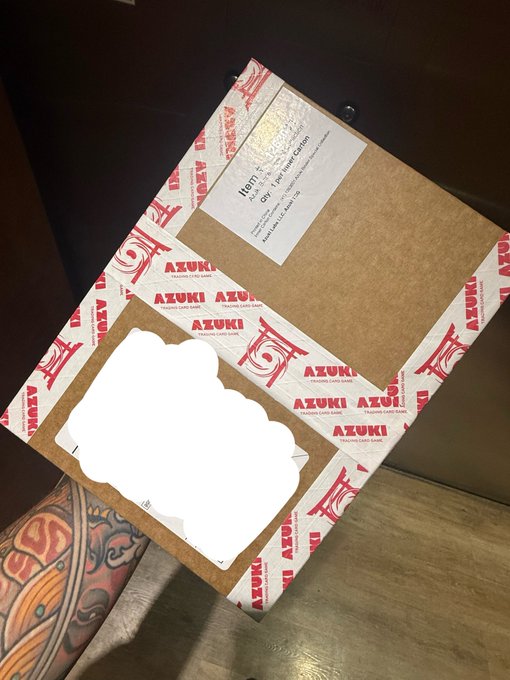 An Azuki trading card game package and a silver-foil 'NOTTE - LIPS LIKE SUGAR' promo card sit on a cardboard backing, next to a pile of white cards and a person's tattooed leg.