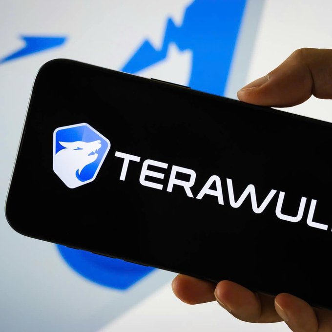 A smartphone displays the "TERAWULF" logo, representing the Bitcoin mining company that has secured a $19 billion AI infrastructure lease deal with Anthropic, driving its stock higher. The logo features a white wolf head inside a blue-and-white shield against a black background, with a blurred blue graphic visible behind the device.