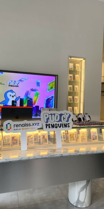 A display setup featuring Pudgy Penguins merchandise, including branded signs and 'renaiss.xyz - Built on Base Chain,' with a TV screen showing a penguin character and shelves of collectible toys in a brightly lit room.